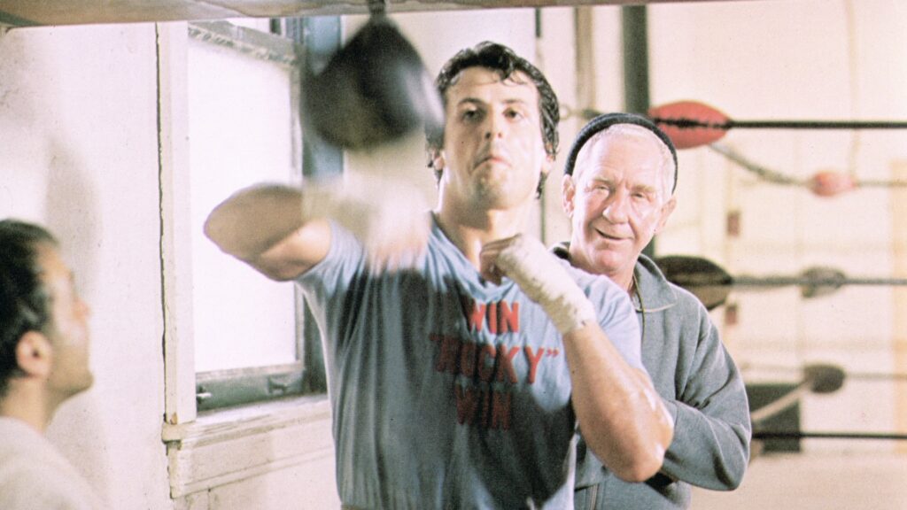 Rocky (Sylvester Stallone) strikes at a punching bag in a boxing ring while his coach (Burgess Meredith) watches during training at a gymnasium in a still from the film, Rocky